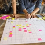 Cafe owner and marketer arranging colored sticky notes on a 90-day desk plan, laptop screen blurred.