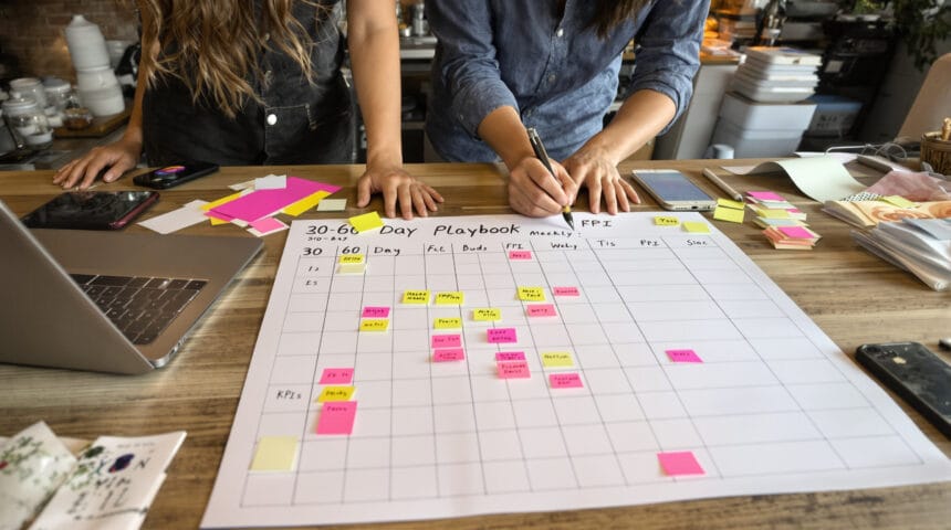 90 Day Lead Nurturing Plan for Small Business: A 30–60–90 Day AI-Augmented Playbook Cafe owner and marketer arranging colored sticky notes on a 90-day desk plan, laptop screen blurred.