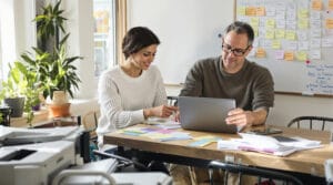 Two small-business teammates vet personalized outreach lines over index cards and blurred printouts at a sunlit wooden table.