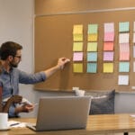 Founder seals an envelope while assistant pins color-coded blank cards into three columns on a corkboard.
