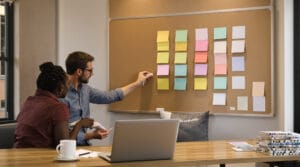 Founder seals an envelope while assistant pins color-coded blank cards into three columns on a corkboard.