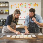Two colleagues arranging color-tabbed index cards into an email-cadence on a wooden table in a roastery back-office.