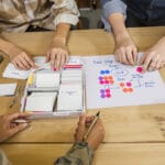 Two-person marketing team sorting plain index cards into color-tabbed trays beside a stickered two-step outreach flow.