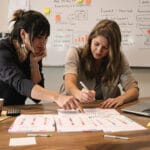 Small B2B team reviewing a blurred spreadsheet and three color-tabbed experiment card stacks at a wooden table.