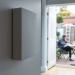 Sharp wall-mounted home battery in foreground; homeowner reviews blurred quotes at kitchen table, EV wallbox visible outside.